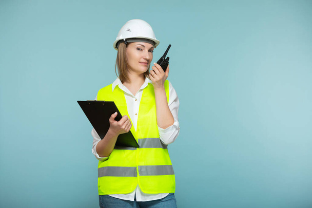 Professional engineer. A woman in a hardhat and bright jelly using a tablet and a walkie-talkie, on a blue background. Copy paste. - Foto, afbeelding