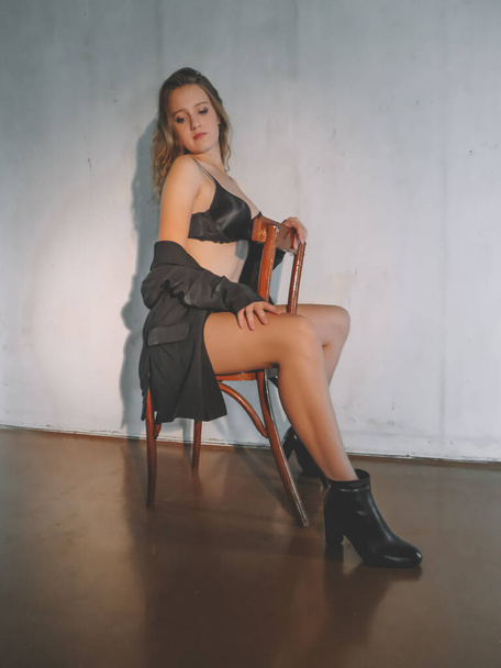 Attractive young blonde woman in black underwear with a slim figure and beautiful legs in boots posing on a chair against the wall, aesthetics and beauty,body care, fashion and style - Foto, Bild