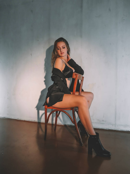 Attractive young blonde woman in black underwear with a slim figure and beautiful legs in boots posing on a chair against the wall, aesthetics and beauty,body care, fashion and style - Photo, Image