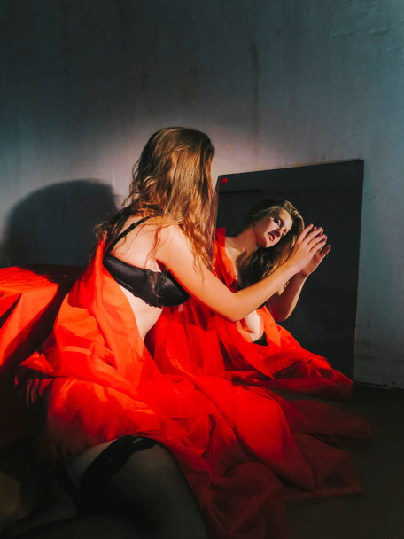 A young beautiful blonde woman in a red veil and black underwear in front of a mirror, aesthetics and beauty of the female body, makeup and cosmetics, body care - Foto, Imagen