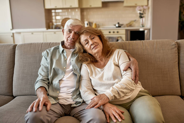 Elderly couple sit on sofa and look at camera - Фото, зображення
