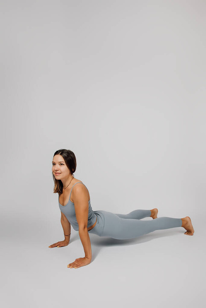 Brunette is doing sports in studio. Young woman in tracksuit performs yoga or stretching exercise, she stands in cobra pose on gray isolated background, side view - 写真・画像