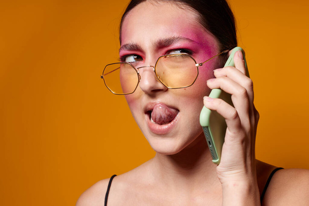 bonita mulher com telefone rosa rosto maquiagem posando atraente olhar óculos amarelo fundo inalterado - Foto, Imagem