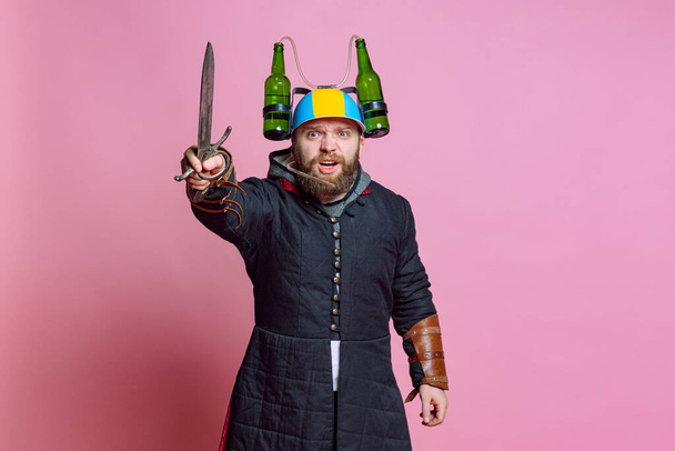Half-length portrait of young bearded man in image of medieval warrior, archer in beer helmet with big knife isolated over pink studio background. Beer festival concept - Fotoğraf, Görsel