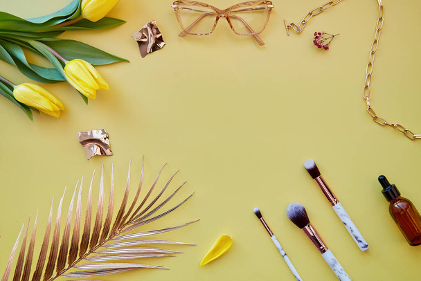 Cosméticos de moda, accesorios, pinceles de maquillaje sobre fondo amarillo pastel. Hojas de palma, tulipanes, gafas, gotero de suero. Copiar espacio - Foto, imagen