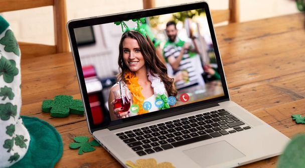 Webcam view of caucasian woman holding a drink on video call on laptop on wooden table. st patricks celebration concept - Photo, Image
