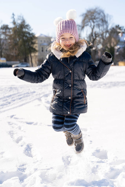 Young smiling preschool girl jumping on snow covered field outside in sunny park - Zdjęcie, obraz