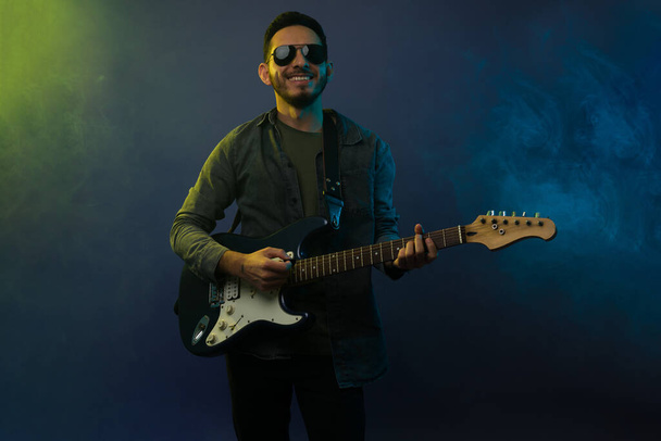 Aufgeregter hispanischer Mann lächelt, während er vor weißem Rauch die E-Gitarre spielt  - Foto, Bild