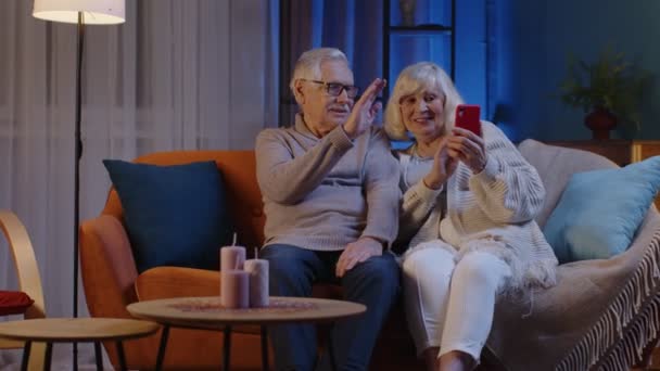 Улыбающаяся пожилая пара бабушка дедушка делает видео-звонок онлайн на мобильном телефоне на домашнем диване - Кадры, видео