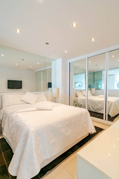 une image d'un lit fraîchement fait à l'intérieur d'une chambre à thème blanche avec une grande armoire en miroir  - Photo, image