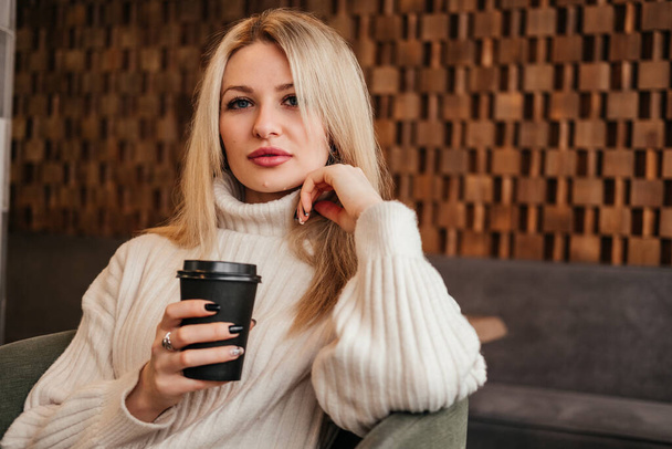 Kobieta w średnim wieku pijąca kawę w kawiarni patrząca w kamerę. biały blondynka dziewczyna w biały sweter wewnątrz. - Zdjęcie, obraz