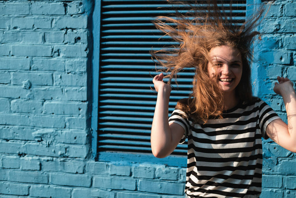 Portrait of a young lovely woman with blue eyes and freckles in a striped T-shirt and jeans against blue brick wall background. - Foto, Bild