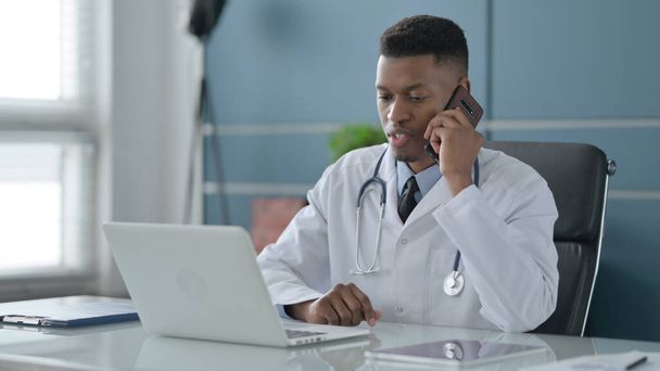 African Doctor Talking on Smartphone while using Laptop in Office - Fotoğraf, Görsel