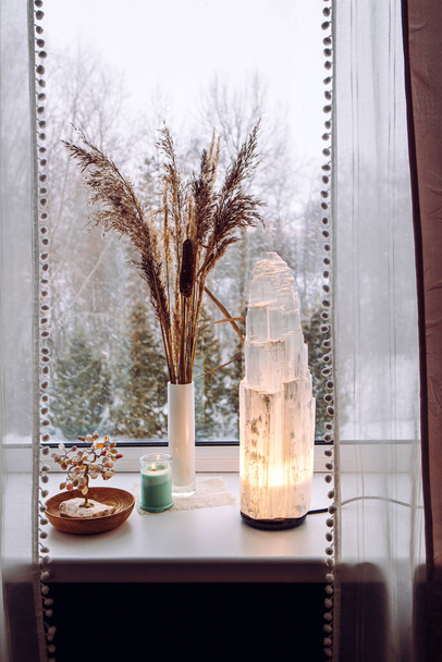 Rough big selenite crystal tower pole lamp illuminated on home window sill, spiritual home decor accent. Winter forest on background. - Photo, Image
