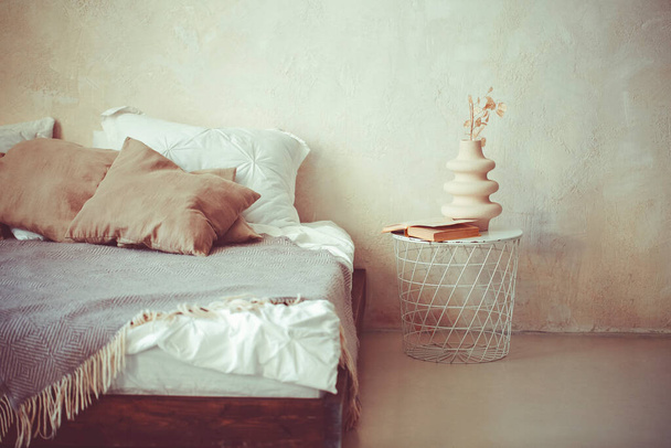 Стильный минималистский стиль лофт спальня интерьер со светлыми бетонными стенами, большая деревянная кровать с подушками, одеяло и металлический круглый прикроватный столик с сушеными цветами в белой керамической вазе - Фото, изображение