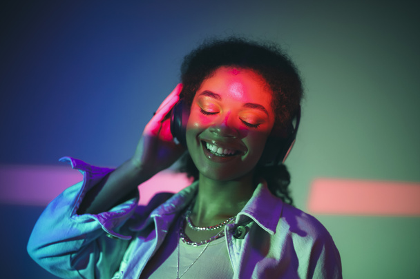 Young happy african american woman in black headphones listening to music, enjoying high quality sound while standing under purple neon glowing light in studio and smiling happily. Musicial equipment - Fotó, kép