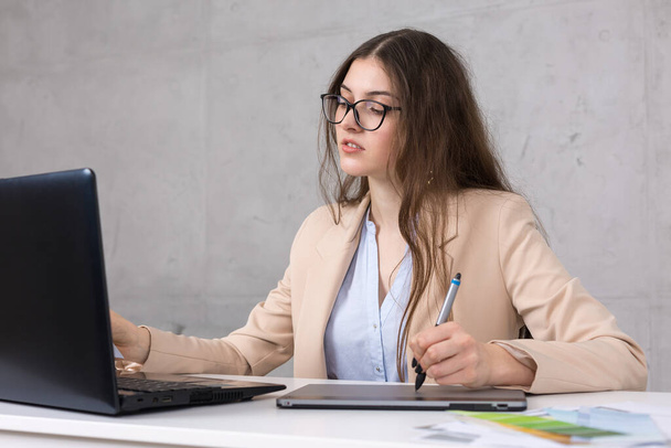 Uma jovem designer sentada em uma mesa desenha em um tablet. Faz cálculos de negócios no computador. - Foto, Imagem