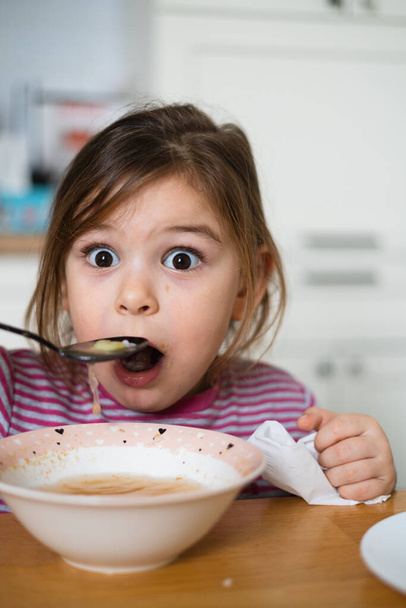 Close up of preschool girl portrait with funny expression eating soup at home  - 写真・画像