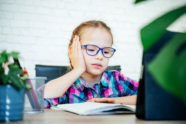 Уставшая маленькая девочка в очках, имеющая онлайн урок, дистанционное обучение для детей - Фото, изображение