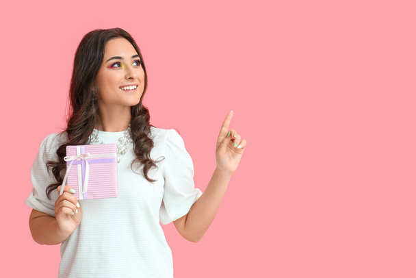 Mujer con maquillaje creativo sosteniendo caja de regalo y apuntando a algo sobre fondo rosa. Día Internacional de la Mujer - Foto, imagen