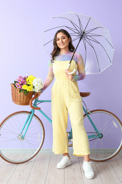 Woman with umbrella near bicycle with basket of flowers on violet background. International Women's Day - Фото, изображение