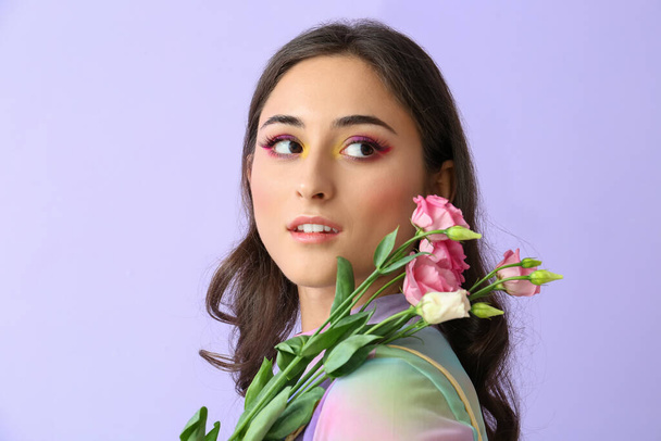 Belle femme avec un maquillage créatif regardant de côté et tenant des fleurs d'eustomie sur fond violet. Journée internationale de la femme - Photo, image
