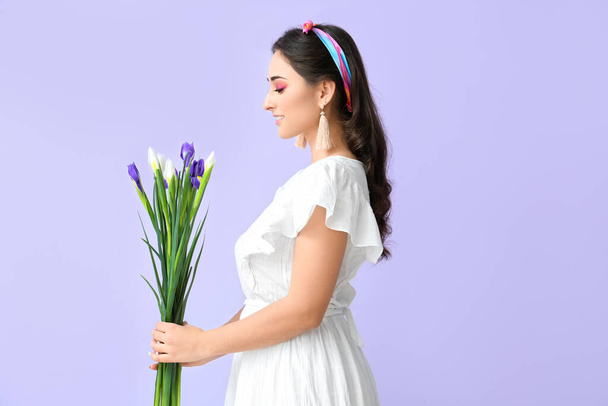 Jonge vrouw met creatieve make-up op zoek naar boeket van iris bloemen op violette achtergrond. Internationale Vrouwendag - Foto, afbeelding