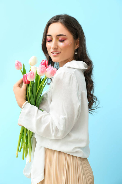 Fashionable young woman with creative makeup holding bouquet of tulips on blue background. International Women's Day - Fotoğraf, Görsel