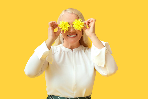 Happy mature woman closing face with flowers on yellow background. International Women's Day celebration - Photo, Image