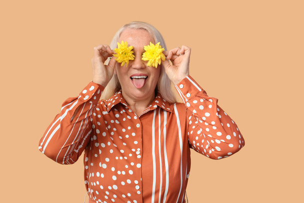 Femme mature avec des fleurs de chrysanthème montrant la langue sur fond beige. Célébration de la Journée internationale de la femme - Photo, image