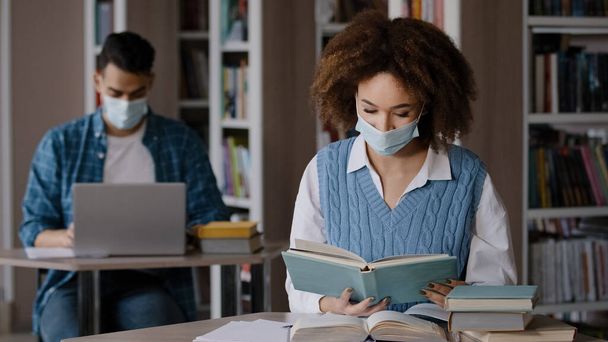 Two students in medical masks studying preparing for exam in university library young guy working typing on laptop girl reading book looking for right information in textbook finds sigh with relief - Foto, Imagen