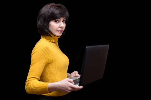 A brunette woman 35-40 years old in a yellow jacket with a high collar with a laptop in her hand on a black background. - Foto, immagini