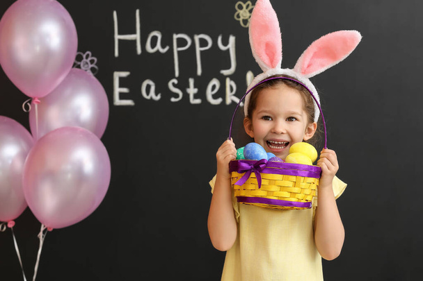 Grappig klein meisje met konijnenoren en Pasen mand op donkere achtergrond - Foto, afbeelding