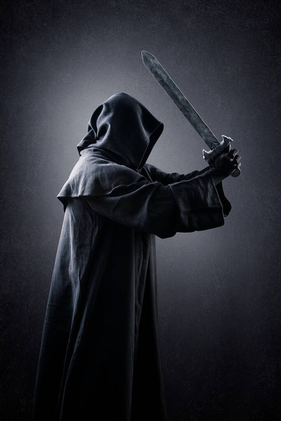 Warrior with hooded cape and medieval sword over dark misty background - 写真・画像