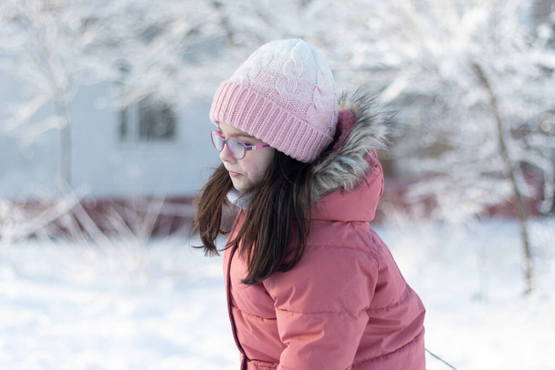Close-up portret van een klein kind in glazen, warme kleren, een gebreide muts en wanten trekt een slee door een touw in de sneeuw. Fotoshoot op een mooie winterse koude sneeuwdag buiten - Foto, afbeelding