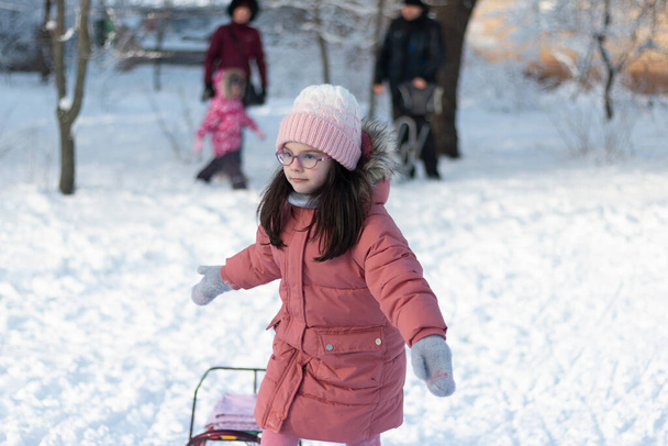 Ένα μικρό παιδί με γυαλιά, ζεστά ρούχα, ένα πλεκτό καπέλο και γάντια τραβάει ένα έλκηθρο από το σχοινί στο χιόνι. Φωτογράφηση σε μια όμορφη χειμωνιάτικη κρύα μέρα σε εξωτερικούς χώρους - Φωτογραφία, εικόνα