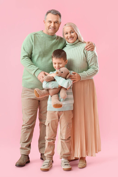 Malý chlapec s hračkou a jeho prarodiče v teplých svetrech na růžovém pozadí - Fotografie, Obrázek