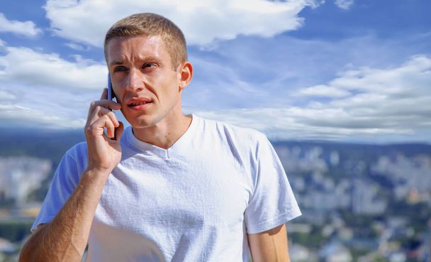 Όμορφος νεαρός επιχειρηματίας κρατώντας gadget και μιλώντας στο τηλέφωνο σε φόντο πανοραμική θέα της πόλης. Προσομοίωση μηνύματος κειμένου.  - Φωτογραφία, εικόνα