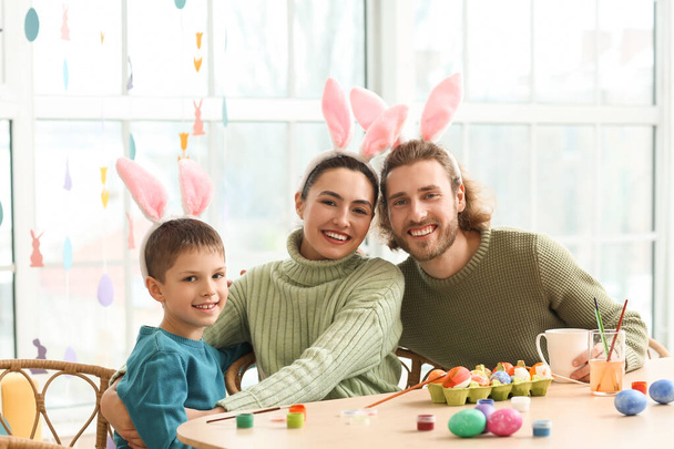 Boldog szülők nyuszifüles kisfiukkal húsvéti tojásokat festenek az asztalnál. - Fotó, kép