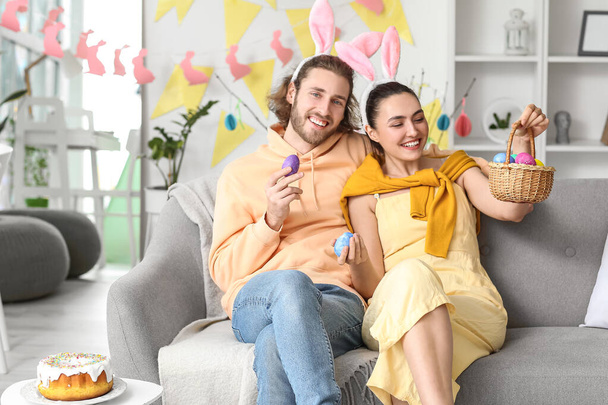 Young couple with basket and Easter eggs sitting on sofa at home - Фото, изображение