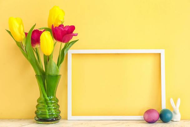 Empty picture frame, vase with tulip flowers and Easter eggs on color background - Foto, Imagem