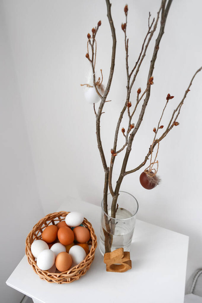 Vase with branches and basket of Easter eggs on table in room - Foto, imagen