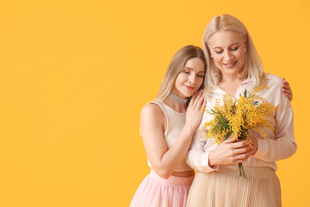 Young woman with her mother and mimosa flowers on yellow background - Photo, Image