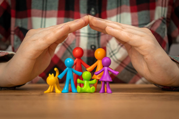 Concepto de seguro familiar con coloridas estatuillas familiares cubiertas por las manos en una mesa de madera. Seguridad y apoyo  - Foto, Imagen