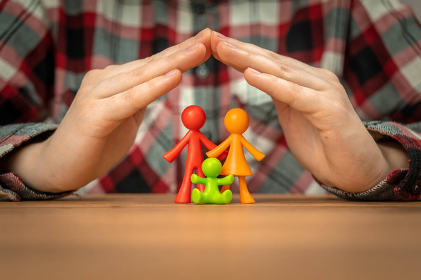 Jeune concept d'assurance familiale avec figurines de famille colorées couvertes par les mains sur une table en bois. Sécurité et soutien - Photo, image