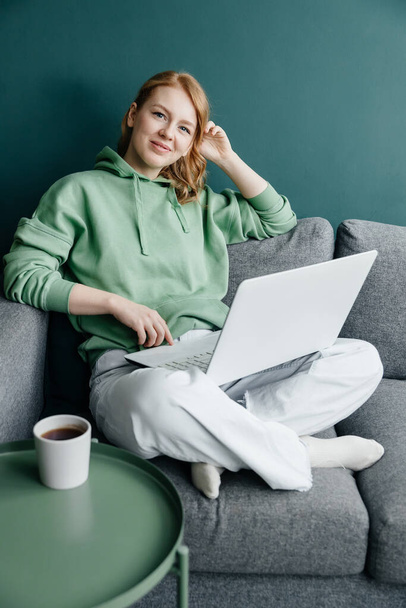 young redhead caucasian woman wearing a green hoodie at home shopping online using laptop - Fotó, kép