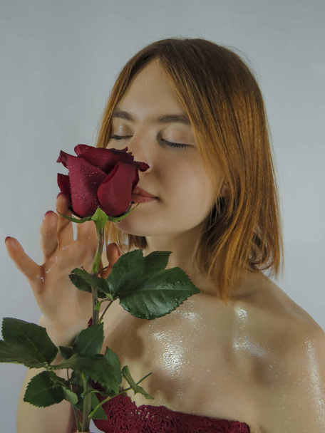 A young brown-haired woman with an attractive appearance in a red negligee with a red rose flower in her hands on a light wall background, close-up, the concept of the holidays of St. Valentine, March 8 - Zdjęcie, obraz