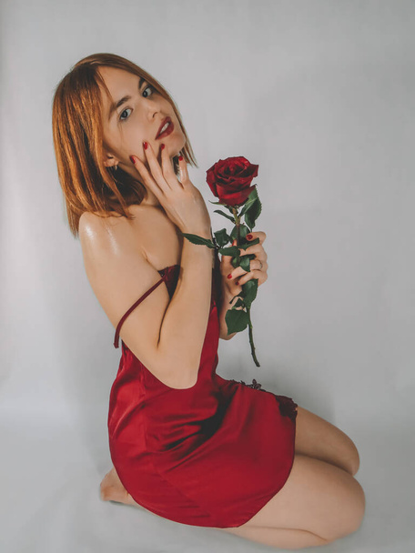 An attractive slender young red-haired woman in a red negligee with a bright manicure on her fingers enjoys the fragrance and beauty of a delicate red rose flower, the concept of a holiday, romantic relationships, attention and care for loved ones - Фото, зображення