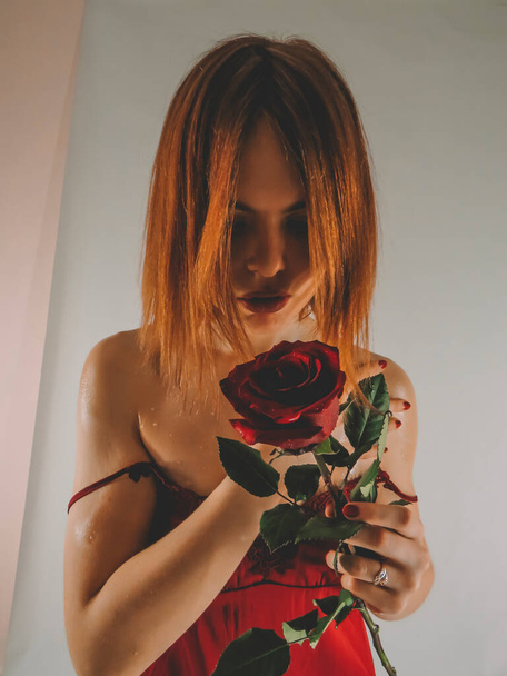 A young brown-haired woman with an attractive appearance in a red negligee with a red rose flower in her hands on a light wall background, close-up, the concept of the holidays of St. Valentine, March 8 - Фото, изображение