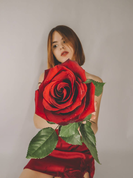 A young brown-haired woman with an attractive appearance in a red negligee with a red rose flower in her hands on a light wall background, close-up, the concept of the holidays of St. Valentine,March 8,signs of attention, romantic dates,female beauty - Foto, imagen